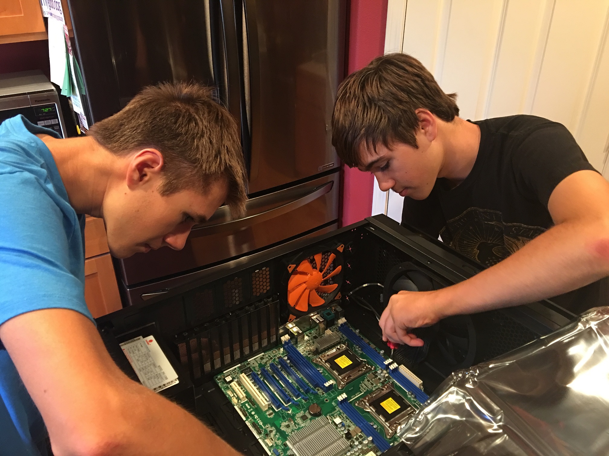 Two teenagers lean over an open computer case, working on a dual-socket server motherboard with two exposed CPU sockets, in a home kitchen.